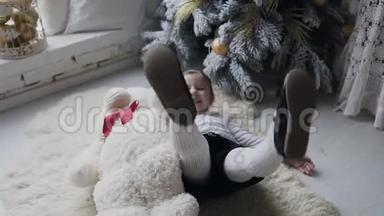 漂亮的宝宝正在客厅圣诞树附近的地板上快乐地玩着一只白色的大玩具熊